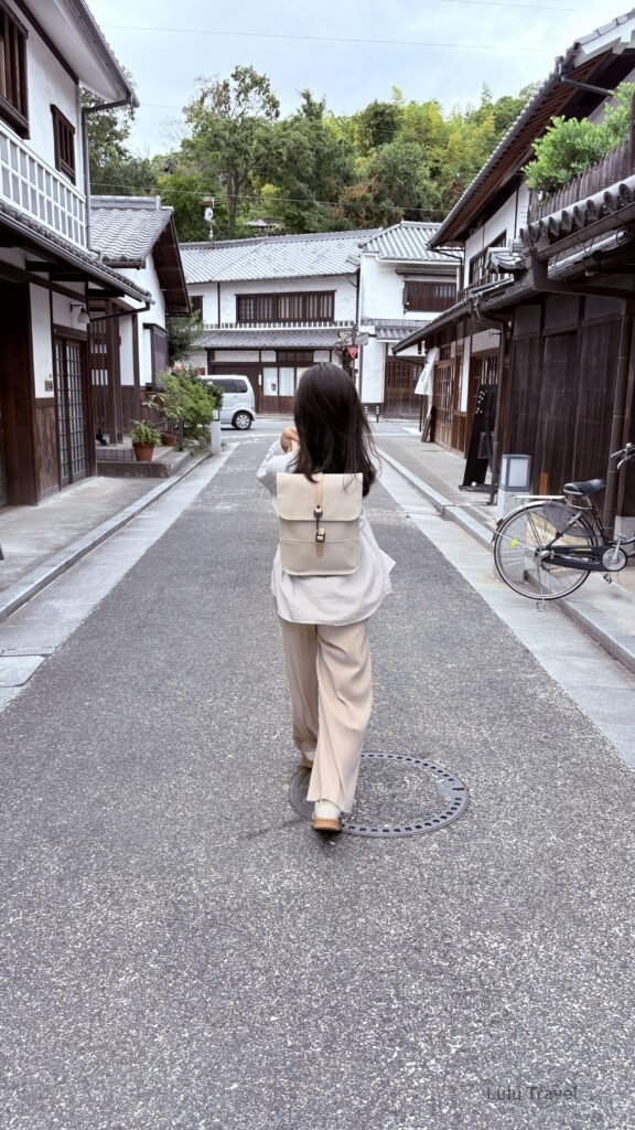 日本傳統老街巷道中，一名旅人背著米色後背包行走在木造町屋之間，兩側是白牆深色木窗的日式建築與停放的腳踏車，街道安靜整潔，呈現慢步調的日本生活風景