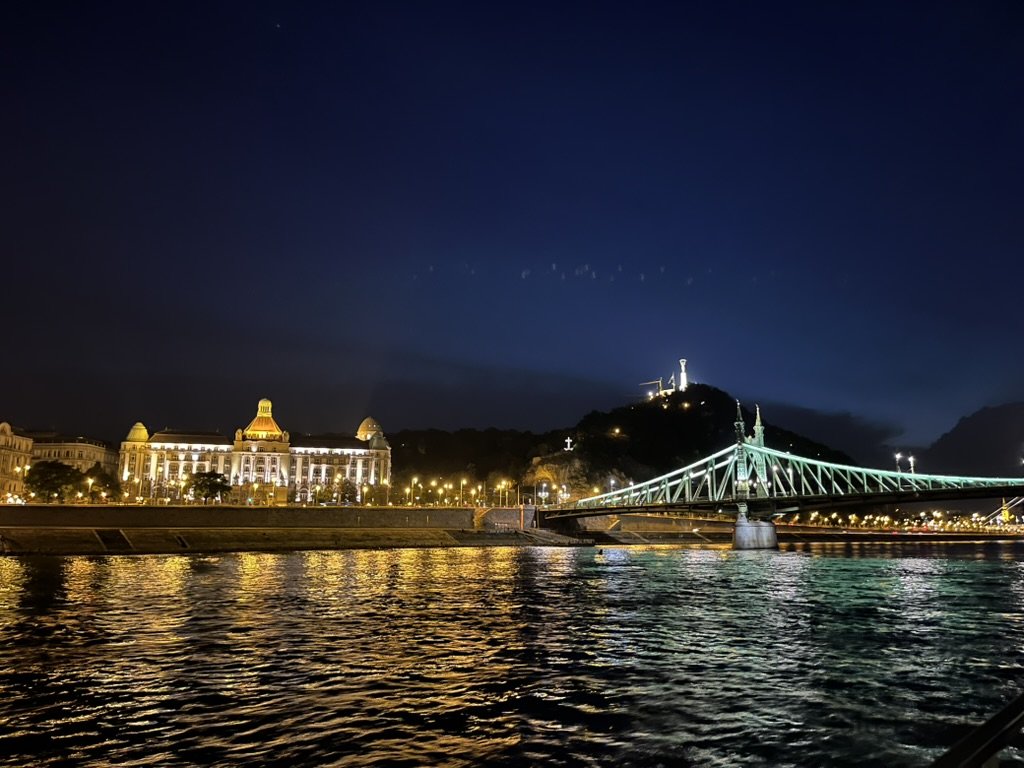 布達佩斯遊船景觀，遠方的自由橋（Liberty Bridge）與蓋勒特丘陵燈火倒映在波光粼粼的水面上。
