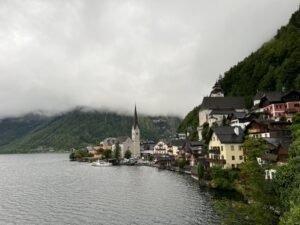 帶長輩去歐洲旅遊|實際安排與心得分享 奧地利哈修塔特湖畔小鎮景色,彩色木屋沿著湖岸排列,教堂尖塔倒映在湖面上,遠方群山被雲霧環繞,呈現雨天的歐洲湖區童話風景