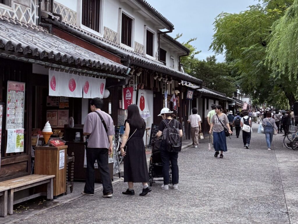 日本岡山倉敷美觀地區，傳統町屋改建的商店街巷道，木造建築與綠意交錯，遊客沿著步道散步