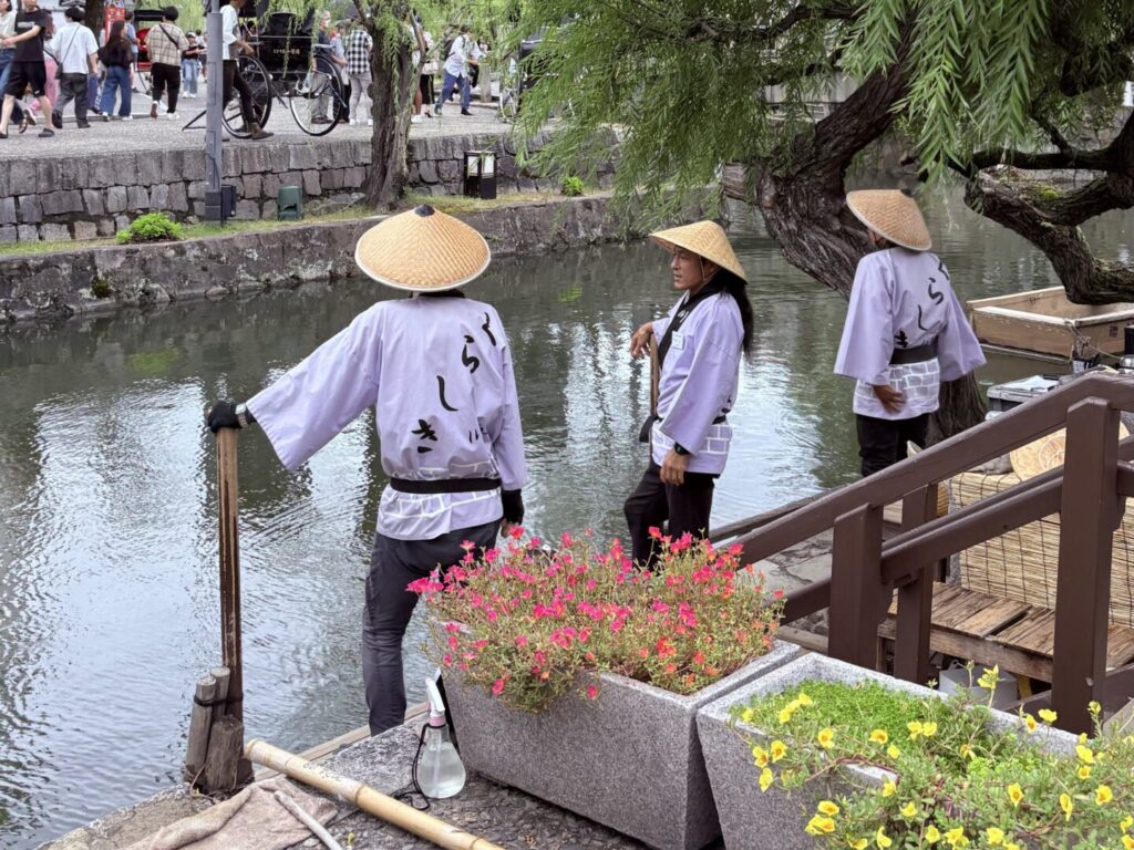 日本岡山倉敷美觀地區，身穿傳統服裝、戴斗笠的船夫在柳樹下準備人力船，河岸旁擺放花盆與竹竿