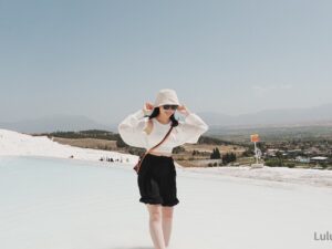 在土耳其白色石灰岩地形前拍攝的旅遊照片，一名女子戴著白色帽子與墨鏡，背景為遼闊山谷與城鎮景色，呈現土耳其獨特的自然與人文風景。