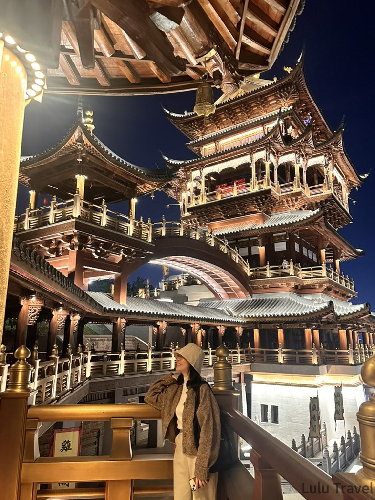 中國出差|深圳的震撼反差與義烏意外挖到的免費景點 在義烏雞鳴閣夜景前拍照留念,戴帽女生站在木構迴廊旁仰望多層傳統古塔,暖色燈光映照整座建築與深藍夜空
