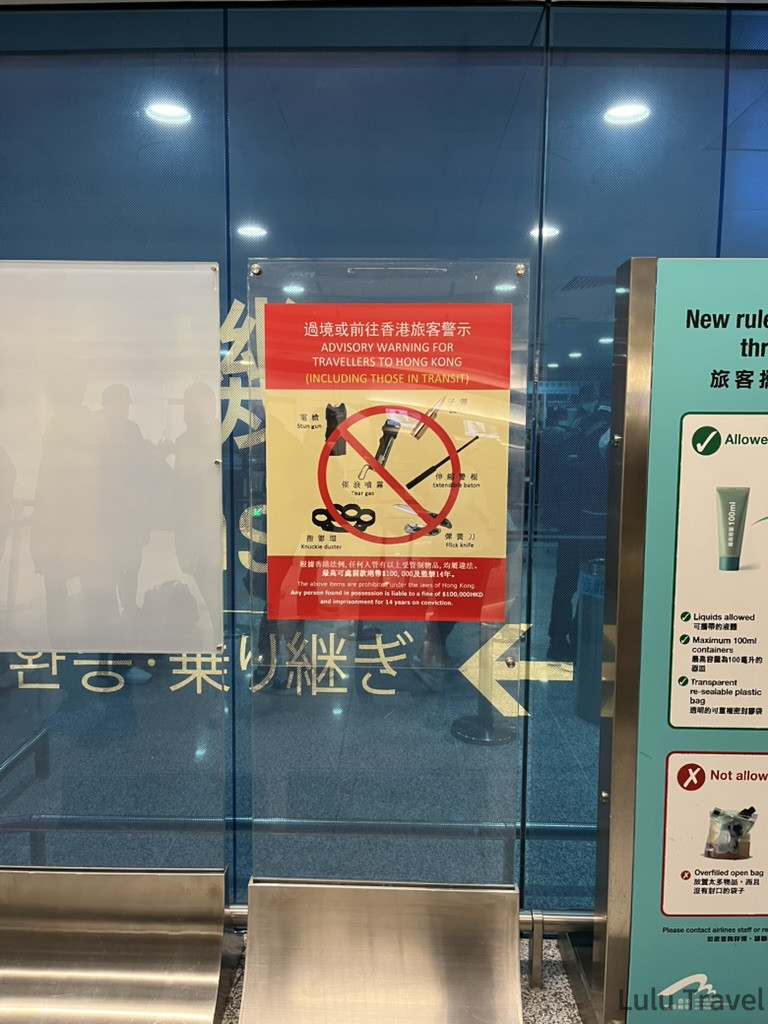 一張張貼在機場透明玻璃隔板上的紅色警告告示，標題為「過境或前往香港旅客警示 (ADVISORY WARNING FOR TRAVELLERS TO HONG KONG)」。告示中央有一個巨大的紅色禁制標誌，列出了香港法律禁止攜帶的違禁品圖像，包括：電槍、催淚噴霧、伸縮警棍、指節環（鐵蓮花）及彈簧刀。 告示下方以中英文說明，任何人管有這些受管制物品即屬違法，最高可處罰款 $100,000 港幣及監禁 14 年。
