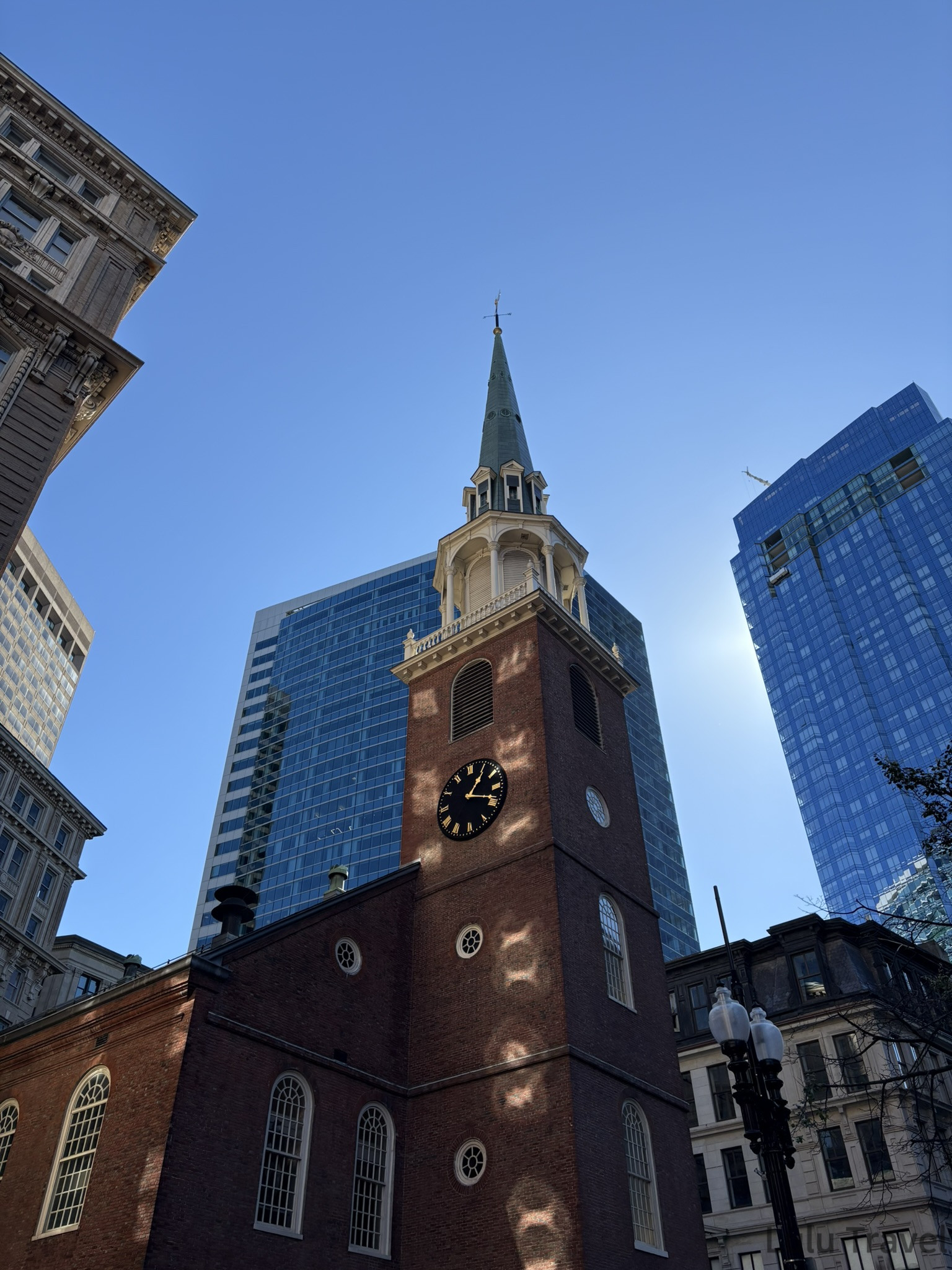 仰角拍攝歷史悠久的紅磚教堂尖塔，南聚會所 (Old South Meeting House)，時鐘指向近四點，背景是現代化的藍色玻璃幕牆大樓，形成強烈的建築對比。