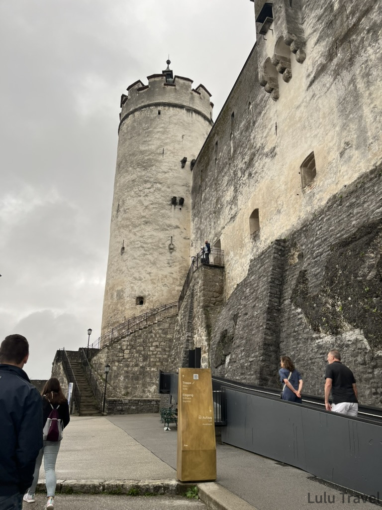 Salzburg Travel Guide — 5 Highlights and Why It's More Than a Mozart Stopover 薩爾斯堡霍亨薩爾斯堡城堡入口,灰色石砌圓形塔樓高聳,遊客沿石階步行進入
