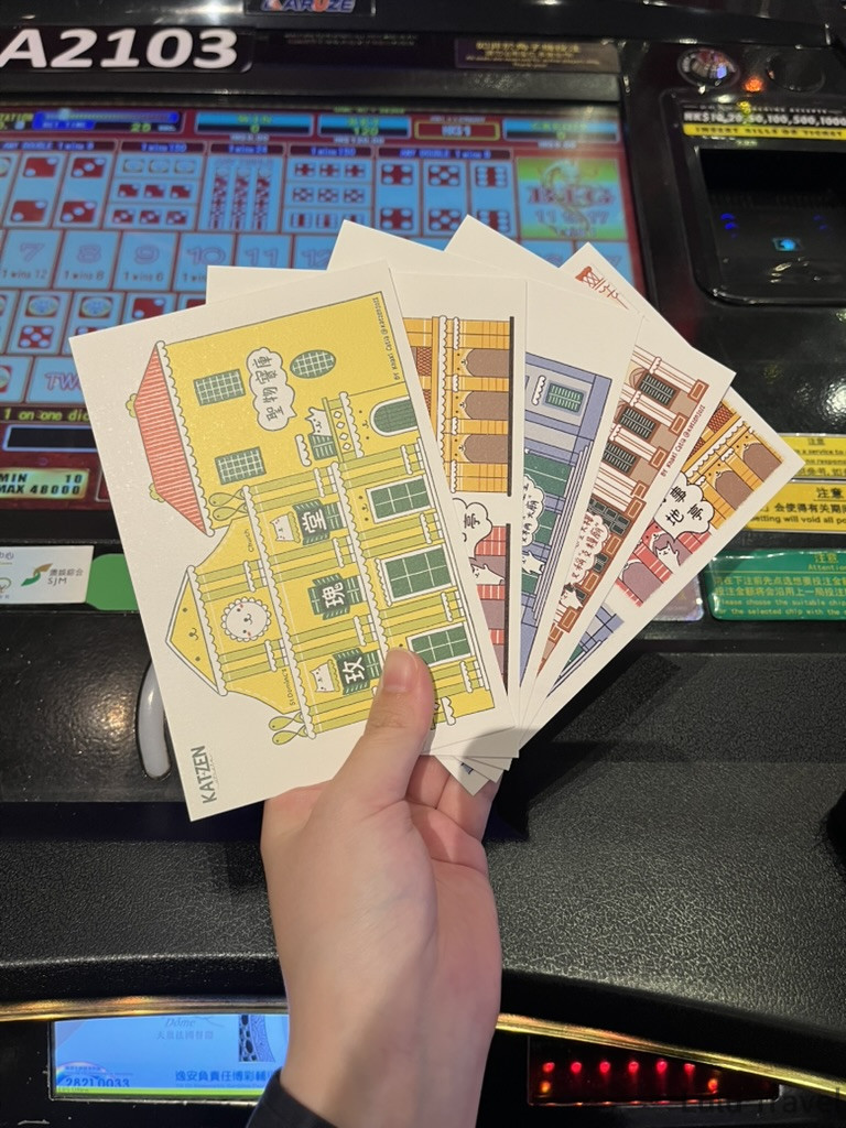 Accidental Macau Travel: A Spontaneous 2-Day Macau Self-Guided Itinerary 在澳門賭場機台前展示葡文書局購入的澳門主題明信片組,背景為彩色電子遊戲機台