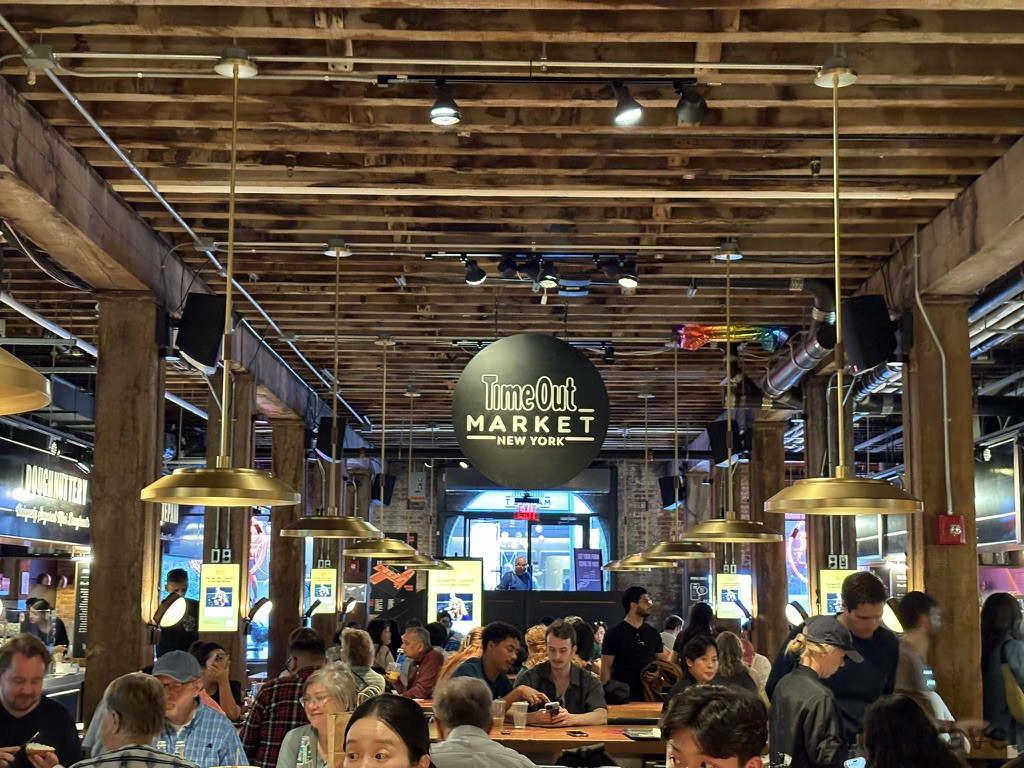 紐約自由行 | 旅費明細、交通技巧與住宿避雷全記錄 紐約 DUMBO 區 Time Out Market 工業風室內用餐區,木質結構搭配工業吊燈與黑色圓形標誌,是 J 人行程中理想的食物中繼站。