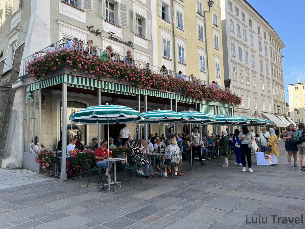 Salzburg Travel Guide — 5 Highlights and Why It's More Than a Mozart Stopover 薩爾斯堡景點 Café Tomaselli 外觀,綠白條紋遮陽棚下坐滿旅客,二樓陽台盛開粉色花卉