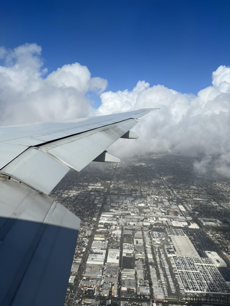 從高空俯瞰城市景觀。機翼的一角佔據了畫面左側。下方是密密麻麻的城鎮建築與街道格線，像是精細的模型。上方則有大片如棉花糖般厚實的白雲，天空呈現深藍色。