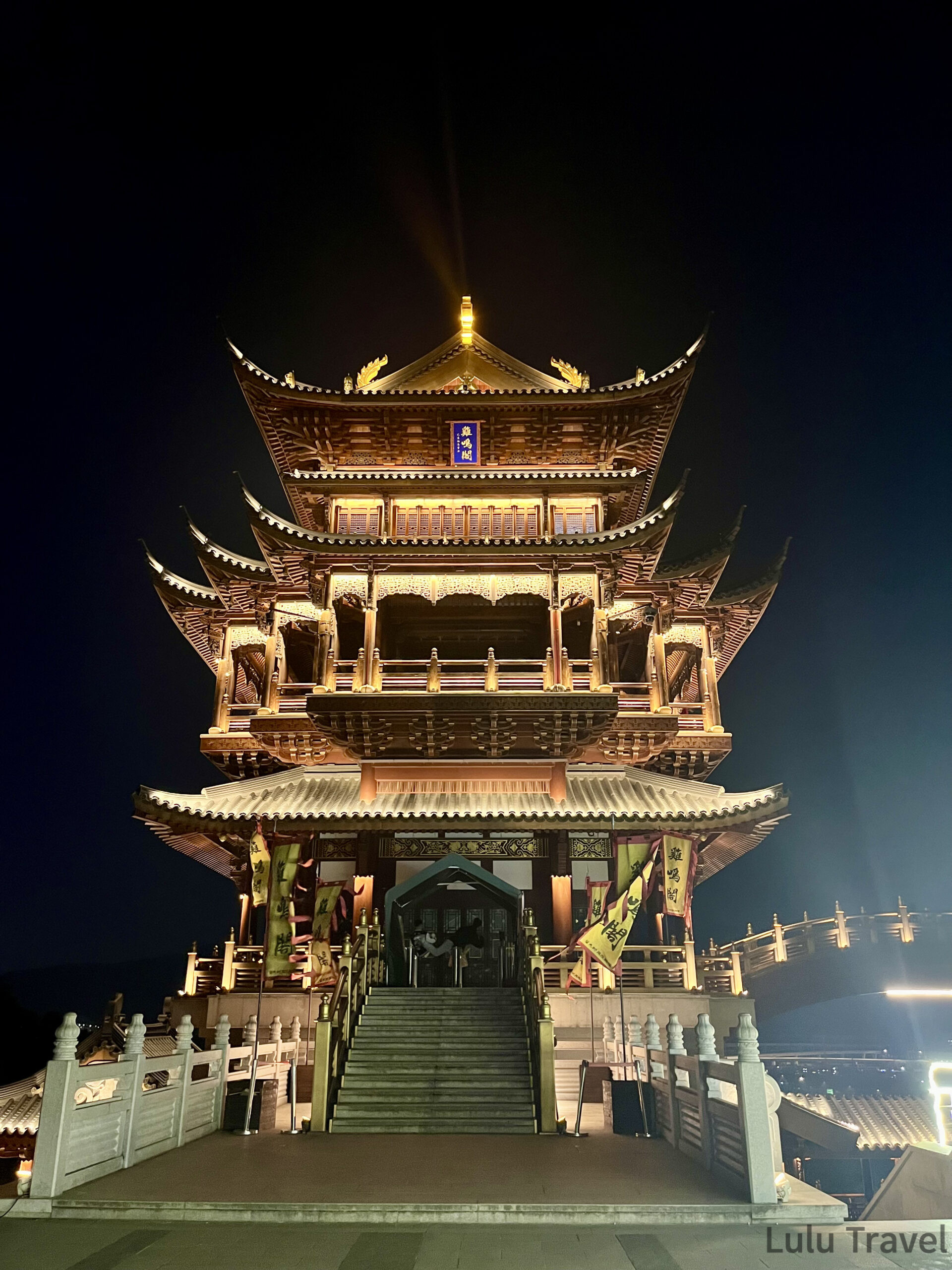 中國出差|深圳的震撼反差與義烏意外挖到的免費景點 義烏雞鳴閣正面夜景,五層中式木構古塔正對鏡頭,頂部金色燈光射向夜空,入口兩側豎立彩旗,石階通往大門