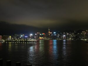 一張在夜間拍下的香港天際線照片。遠處的摩天大樓在夜色中發出閃爍的光芒，維多利亞港的水面也倒映著這些燈光。天空陰雲密布，右側隱約可見山脈。左側水面上有一艘渡輪，渡輪上的燈光格外明亮。