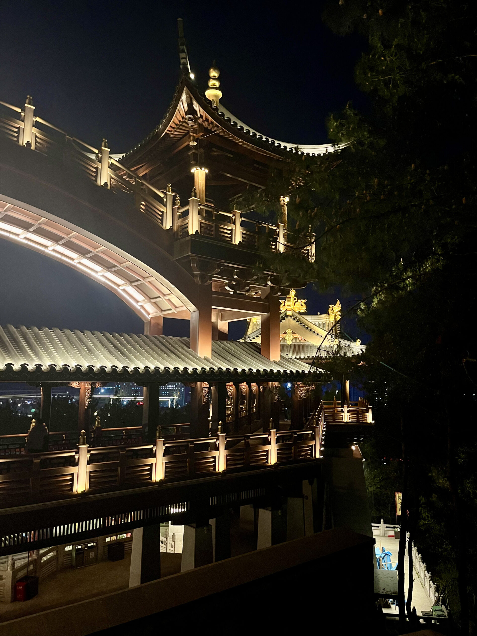 中國出差|深圳的震撼反差與義烏意外挖到的免費景點 義烏雞鳴閣夜間側面建築,傳統中式木構飛簷與弧形拱橋在暖色燈光映照下,背景可見義烏市區夜景