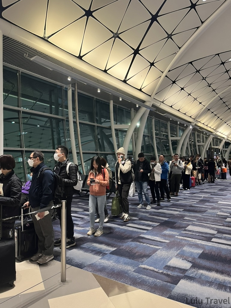 香港機場夜間旅客排隊人潮，延伸至幾何網格天花板下的長廊，出差回程趕班機的現場