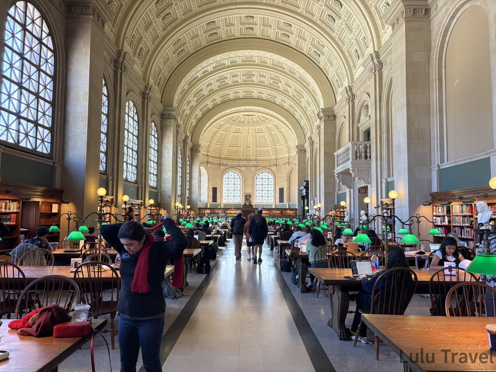 Boston Day Trip from New York — Is 4 Hours Each Way Worth It? 波士頓公共圖書館 (Bates Hall)壯闊的巴洛克式拱頂閱讀室。長型木桌上擺放著標誌性的綠色罩檯燈,兩旁是整齊的高聳書架,讀者在靜謐、古典的氛圍中專注學習。