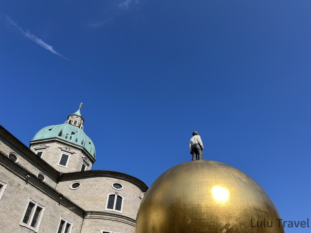 Salzburg Travel Guide — 5 Highlights and Why It's More Than a Mozart Stopover 薩爾斯堡主教座堂旁的金色球體雕塑,球頂站立小人像,背景為教堂翠綠圓頂與晴天藍空