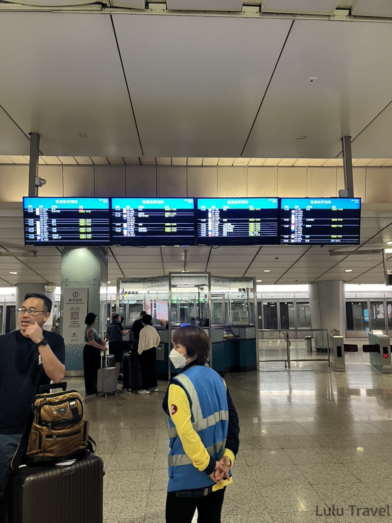 一張在香港機場快線站外拍下的照片。有一位穿著藍色反光背心的志工，在現場指引。身後可以看到數塊顯示班次的螢幕，下方則是幾位正等待購票的乘客。