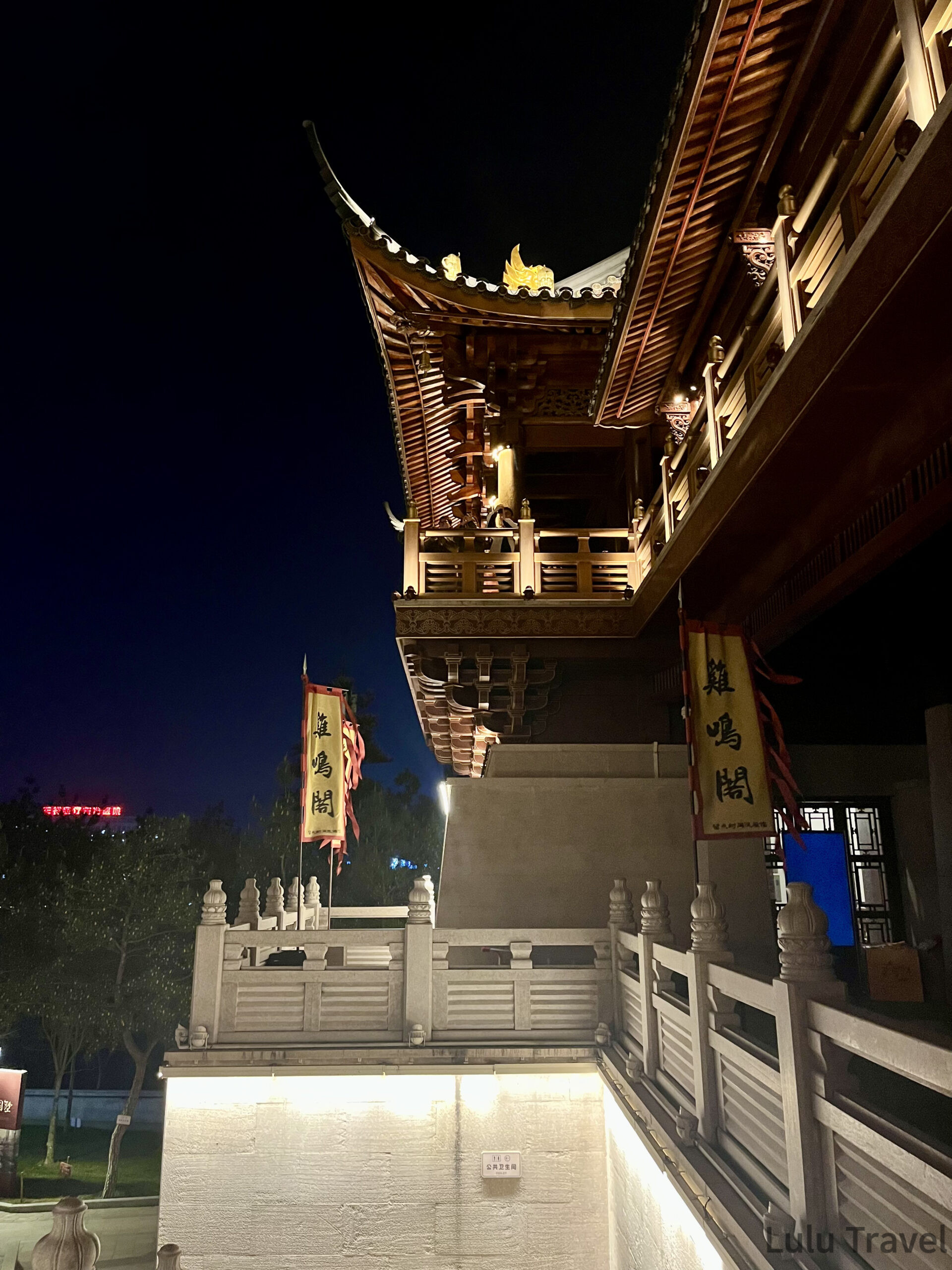 中國出差|深圳的震撼反差與義烏意外挖到的免費景點 義烏雞鳴閣近景仰拍,中式飛簷屋頂與木構迴廊在夜燈下顯現精細雕刻,前景為白色石欄與雞鳴閣旗幟