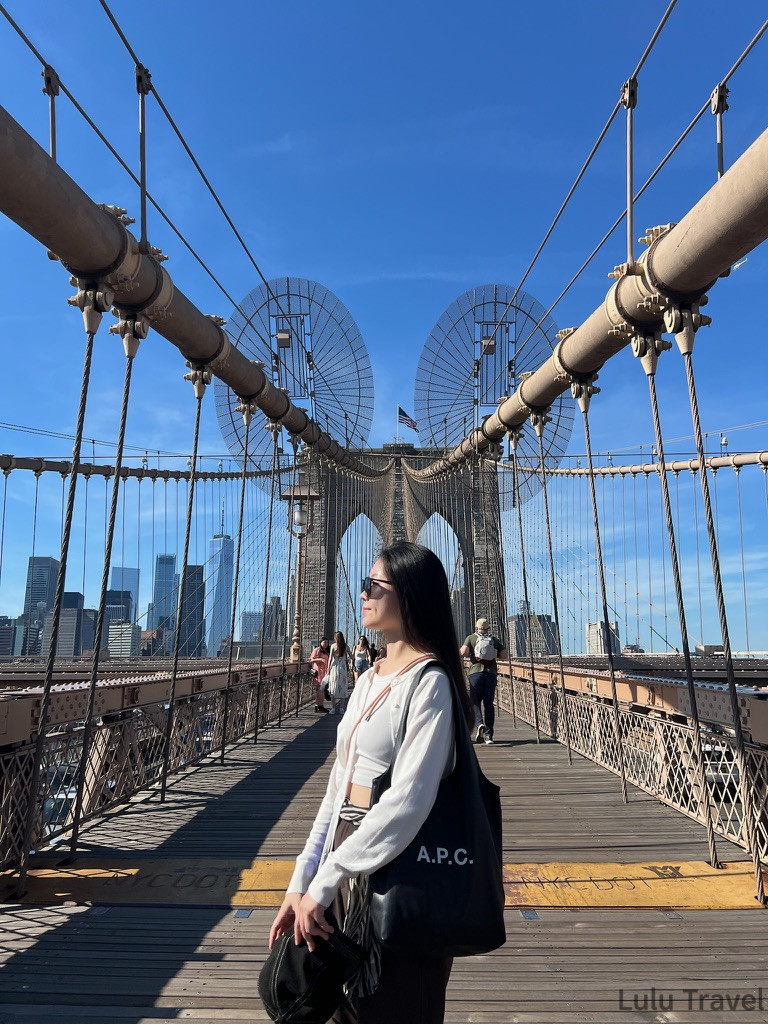 紐約行程規劃|DUMBO、百老匯、布魯克林到波士頓一日遊 旅客穿著白色針織上衣、背著 A.P.C. 帆布袋走在布魯克林大橋(Brooklyn Bridge)木質步道,背景為湛藍天空與大橋鋼索結構。