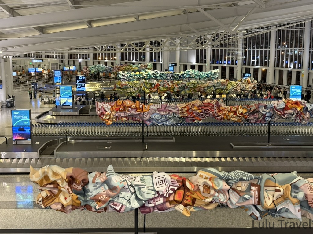 紐約自由行 | 旅費明細、交通技巧與住宿避雷全記錄 西雅圖機場(SEA-TAC)行李領取大廳實景,行李轉盤上方有色彩鮮艷的藝術裝置。