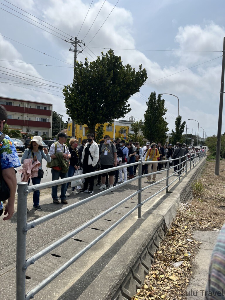 日本郵輪旅行 | 高雄出發歌詩達莎倫娜號的 10 個真實心得 戶外街道旁的一群旅客正沿著金屬護欄排隊步行,背景可見低矮民宅、黃色建築與綠樹,天氣晴朗。