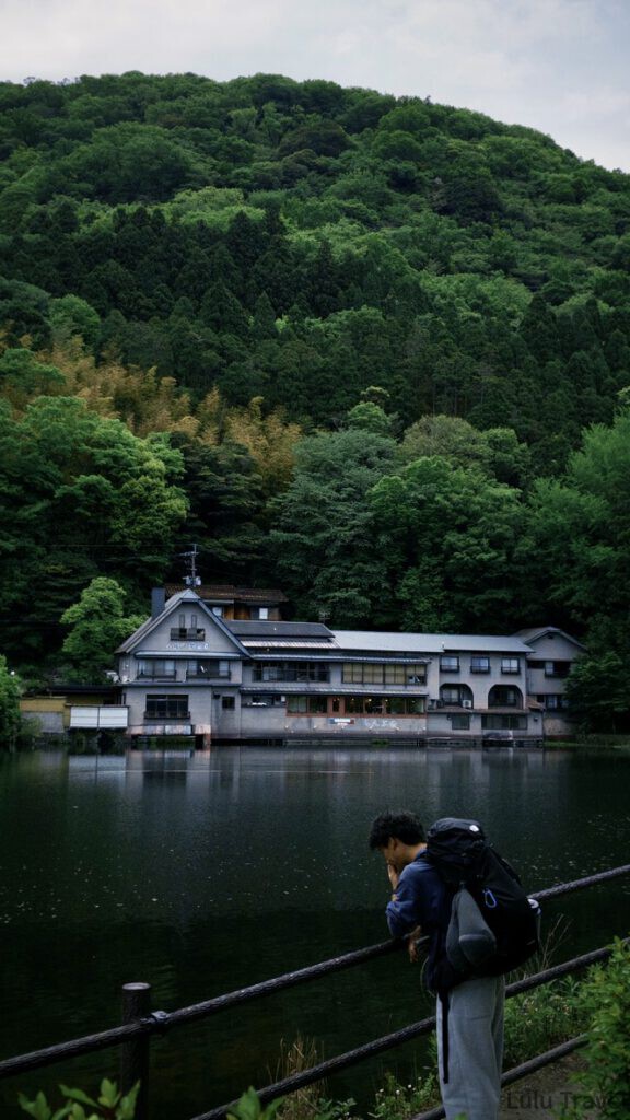福岡自由行|日本黃金週的6 天 5 夜行程與消費明細 清晨的金鱗湖,湖面如鏡倒映著周圍茂密的綠色森林與湖畔傳統日式建築,一名背包客正倚靠在欄杆上欣賞美景