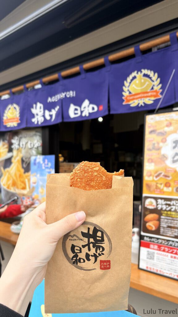 由布院兩天一夜|避開黃金週人潮的溫泉住宿與由布院之森 手持揚げ日和的金賞咖哩麵包,背景是店家的藍色暖簾與咖哩麵包大獎看板